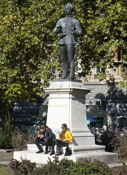 Twee mensen zitten op de sokkel van een standbeeld. Alle drie kijken op hun telefoon.