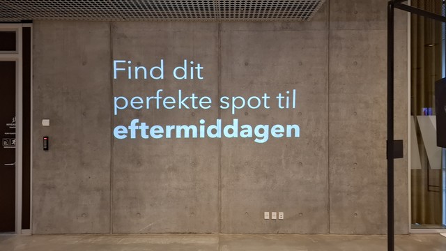 Projected in turquoise letters on a concrete wall inside Dokk1 Aarhus main library, the sentence: Find dit perfekte spot til eftermiddagen. 'Find the perfext spot for the afternoon.' 