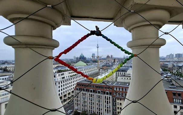 Looking across Berlin from a height of 50 metres. We are on the observation deck of Französischer Dom - the French cathedral. The balustrade is fenced off with a metal security net that makes a pattern of  diamond-shaped openings. Someone has wound multi-coloured threads round one little diamond to create a rainbow border. We are looking through the rainbow diamond as it frames two of the highest and most famous landmarks, tiny in the distance - Fernsehturm (television tower) and Berlin Domkirke (cathedral). Like my hometown of Aarhus, Berlin is one of the official Rainbow Cities in the Rainbow City Network that works for social inclusion and LGBTI+ rights, so this rainbow framing feels right.