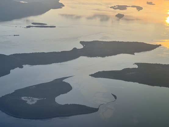 An arial sunset view of islands off the southwest coast of mainland Canada. The waters are  deep blue near the bottom of the photo and transition through orange to yellow near the top right corner. There are also clouds reflected in the ocean near the top of the image. The islands have irregular shapes, including a heart-shaped pond in the bottom-most island [April 2024] 