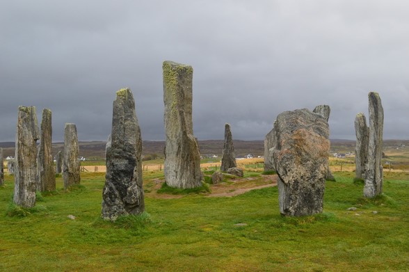 A large stone circle - the central circle at Callanish - with one noticeably taller stone in the centre. The sky is grey and overcast, the landscape flat and bleak. The stones are blunt-edged and textured, patterned with with lichen and grooves.