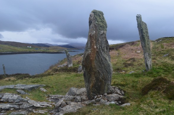 A semi circle of upright stones are shown on the edge of a loch, in a rough field of grass and rocks. Beyond a narrow straight of water, there's the low profile of Lewis under some dark clouds, a white croft visible in the distance. The stones are tall and ridged.