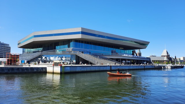 Looking from an outer pier at Aarhus Harbour in towards Dokk1 – Number 1 Dock. This is both the name of the main dock the mid-city port and the name of the library that was built on it about 8 years ago. The library is a huge, striking, four-storey modern geometric building right on the waterfront with picture windows and  wraparound terraces and outdoor playgrounds and seating areas and community spaces that maximise the fabulous view and location. The sky is cloudless and light blue. Beyond the city skyline including spires and turrets on historic buildings. Symbols for the 17 UN Sustainable Development Goals are painted in white on a lower section of the library. The sea in front of it is rippling, deep blue. In a splash of contrasting colour, a bright orange dinghy is just sailing past. We can just see the tiny figures of kids playing and people walking, cycling, sitting, hanging out on the terraces.
