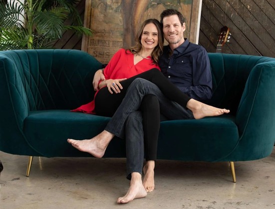 Gia Mora and Denny Rivas sit on a couch with their legs crossed over each other, smiling at the camera