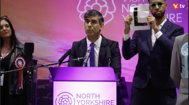 Moment from Rishi Sunak's speech at the results announcement for Richmond and Northallerton. He's shown at an illuminated 'North Yorkshire' lectern in white and purple. Behind Sunak, a tall man - the YouTuber Niko Omilana - is wearing a pair of small sunglasses and holding up a white sign with a large black 'L'.