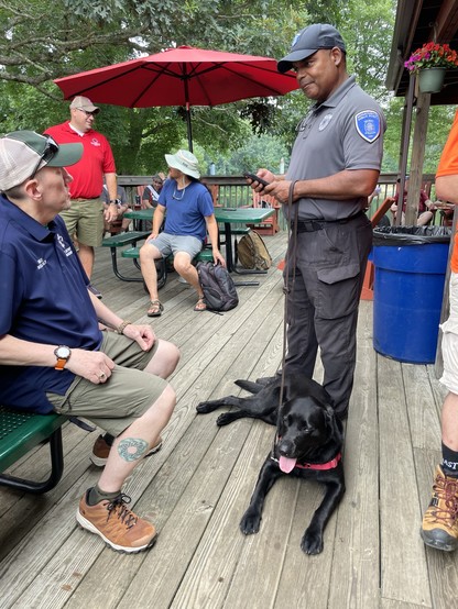 An officer from the University of Hartford talked to a person seated while his dog, a black Labrador rests on the deck at his feet. 