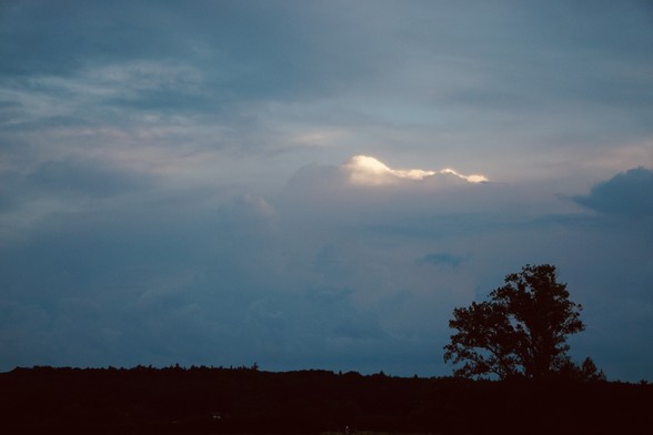 A serene landscape with a dark silhouette of trees in the foreground and a vast sky with moody clouds at twilight. There is a small patch of light breaking through the clouds.