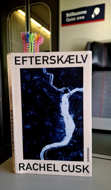 My Danish-language paperback library edition of Rachel Cusk's Aftershock. 'Efterskælv.'  Sans serif slim black typeface. Broad off-white border. Above: title Efterskælv. Below: author name. Between them: big bland navy-blue image. I think it's the ocean with a thick white fault-line of white foam snaking through it. 