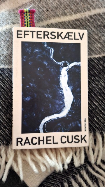  My Danish-language paperback library edition of Rachel Cusk's Aftermath. 'Efterskælv.' Sans serif slim black typeface. Broad off-white border. Above: title Efterskælv. Below: author name. Between them: big bland navy-blue image. I think it's the ocean with a thick white fault-line of white foam snaking through it. There's a pink and yellow woven bookmark sticking out more than halfway through the 200 pages. The book is lying on a wool blanket, because you need a blanket and a hot water bottle for reading on the couch in Denmark in the middle of Summer.