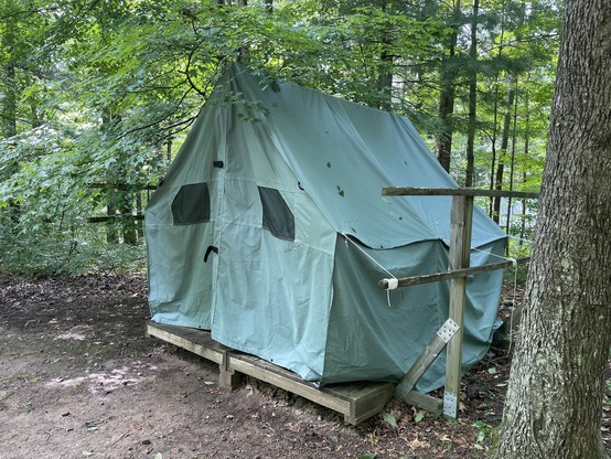 A large green canvas tent on a wooden platform. The screened windows look like eyes and a black strap looks like a little sad mouth. 