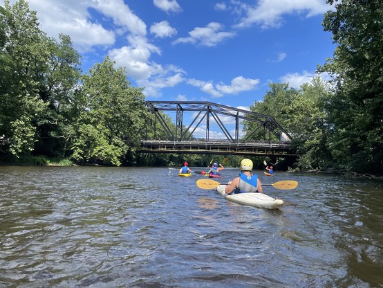 Several people kayaking on a river under a bridge, surrounded by lush green trees on a sunny day. Farmington River, Farmington CT, US. 