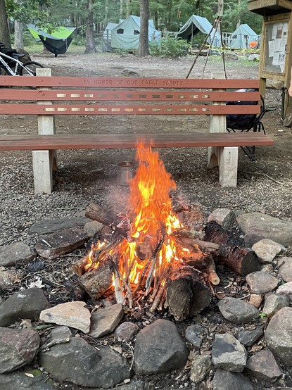 A campfire surrounded by large rocks with a wooden bench in the background that reads 