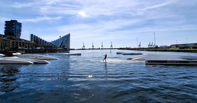 Water’s edge, Aarhus city centre, looking out to sea. Left: distinctive triangular and rectangular and octagonal apartment blocks, including a 70 metre tall one with a giant clock. Right, across the water: the docks with warehouses and industrial cranes like giant’s chairs. In the expanse of water in front of us, gleaming white water-sports ramps below cables. In the middle, a single person in a wetsuit, upright at full speed on a wakeboard, a trail of bright white wake behind them. The sky and sea are deep blues, the sun is very strong, sparkling on the water. The whole scene is extremely blue. After months of wind and winter-grey summer days and incessant rain, today feels like stepping through a portal into a parallel universe where everything is made of this amazing newfangled colour, blue. 