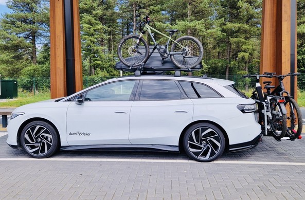 White electric car with two bicycles mounted on the rear rack and one bicycle mounted on a roof rack, parked in a forested area.