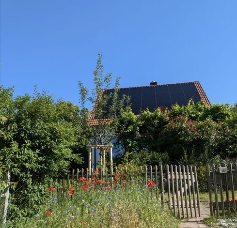 Man sieht einen Garten, Blumen und ein Gartentor. Im Hintergrund ein Haus mit Solaranlage, dahinter blauer Himmel