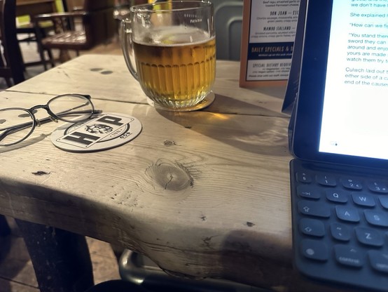 A pint of Budvar, a pair of glasses and an iPad with a keyboard attached on a wooden bar table, a beer mat says Hop Inn