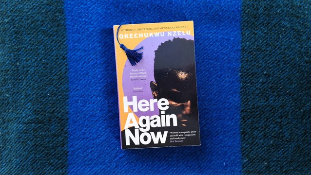 My paperback edition of Here Again Now by Okechukwu Nzelu. A glossy, striking cover with a distinctive mustard-yellow and purple background and a close-up portrait of a young man's face - the protagonist, a Nigerian-English man. There are praise quotes from Derek Owusu; Brit Bennett; and The Guardian. The book is lying on a wool throw-blanket in deep blue shades. My bookmark is sticking out about 30 pages in - it has a blue silk seventies-curtain-style tassel that I love in a not even slightly ironic way.