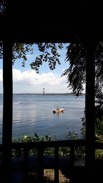 Upright image. Sitting in a gazebo on a hill in Middelfart looking across the bay. The scene is framed by the wooden posts of the gazebo and leafy tree branches outside. Between them, we see calmly rippling blue sea below a light blue sky with fluffy white clouds. Alone on the water: a single orange, red and white fishing boat. Beyond: Lillebæltsbroen. The Little Belt Bridge. The 1,700 metre long, 60-metre high suspension bridge that links the island of Fyn (Funen) to the Danish mainland of Jylland (Jutland). We can see a tree-lined sliver of land to the right where the bridge ends. Here Be Jutland. 