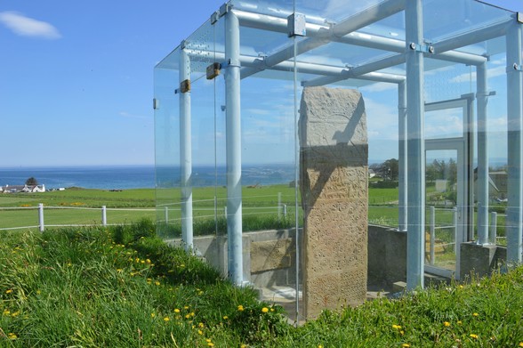 A 2.7m standing stone is encased in a glass 'cage'. The stone is a dusty pink and there are intricate symbols carved into it. It's isolated in a large green field - the sea blue and calm in the distance, the sky clear.