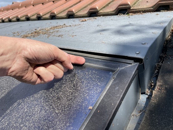 A close-up of a hand lifting the edge of a metal panel on a roof, revealing a gap. The roof has both metal and tiled sections, with some debris visible.
