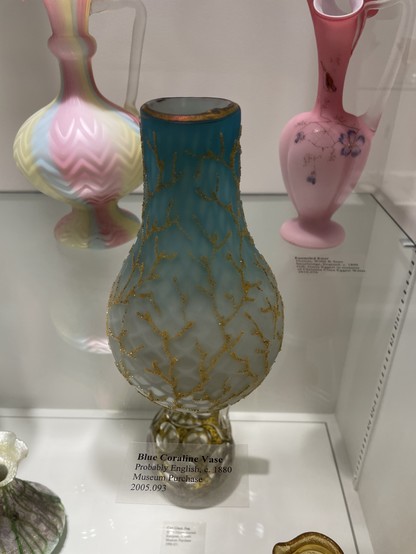 A clear glass shelf at the New Bedford Glass Museum with a blue vase with a raised gold coral like pattern possibly from England 1880. Behind are two other opaque vases, one rosy pink with a floral design, the other with blue yellow and pink stripes. All three vases, while different, have larger bulbous bottoms and thin, narrow tops. The blue and pink vases are darker on top and light on top. 