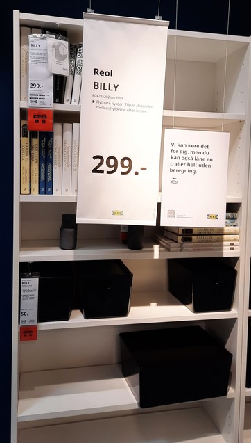 Bad blurry image of a Billy Bookcase on display in a dark Ikea showroom. 2 metres high, simple, white. There are some ornamental books on it. There are (Danish-language) signs hanging off it with the specifications, price 299 kr. (about 40 euros) and delivery information.

