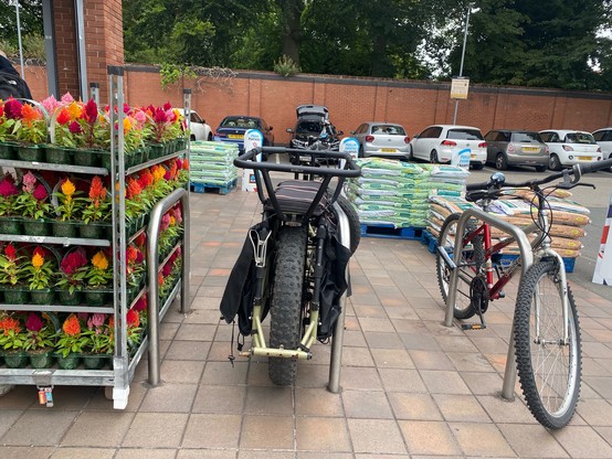 A set of 3 bike stands outside a supermarket. One stand is obstructed by a trolley of plants. Wooden pallets with big bags of soil are scattered around the pedestrian entrance.