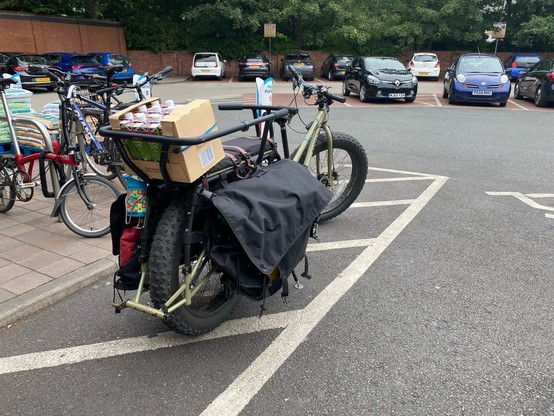 A longtail cargo bike loaded up with shopping. Bulging panniers to the sides and a big box of milk cartons bungeed to the rear child seat.