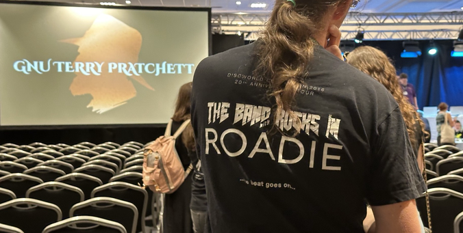 A man wearing a tshirt which says on the back ‘The Band With Rocks In ROADIE’ and ‘Discworld Convention 2016’. A screen says ‘G.N.U. Terry Pratchett’ in the background. I am wearing the ‘Band With Rocks In’ tshirt from Discworld Emporium.