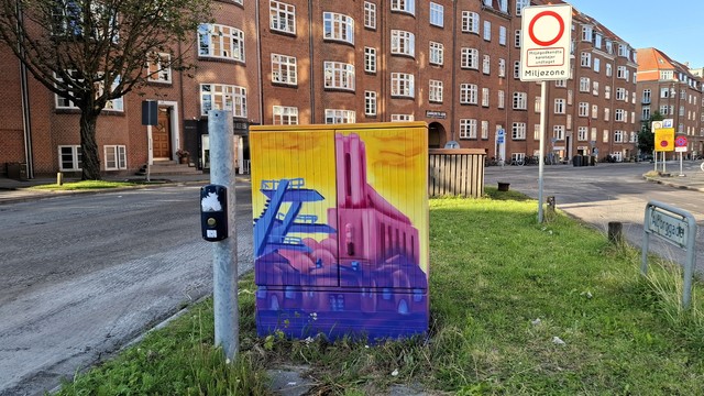 Triangle between roads that meet in Frederiksbjerg, a mid-city mixed-use neighbourhood, Aarhus.  Beyond: picturesque centuries-old six-storey terraced apartment blocks characteristic of this area. We can see bike paths, parked bikes, residential rubbish-sorting stations, traffic signs. All the usual city centre paraphernalia.  On this triangular verge: a pedestrian traffic light button and a street name sign: Ålborggade. Central to it all, popping with colour: a utility box painted in vivid deep yellows, pinks and blues. The artwork is local landmarks. Diving platforms at the nearby swimming arena. The unique shape of nearby Sankt Lukas, a 1920s church shaped as a 48-metre stone rectangle that's topped by a thick 35-metre oblong tower. Now follows same context description for all photos in this thread, if you want to skip. A council utility box for electrical and technical stuff, about half a metre by half a metre in size, normally grey metal box, typically dirty and covered in graffiti and stickers. They have been transformed all across the city in recent weeks, painted on all four sides and on top with images that reflect something in the immediate neighbourhood. Artist signature: J W ’24.