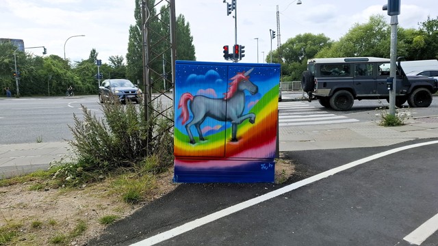 Ringroad crossroads, Aarhus. Trees and traffic lights and an electricity pylon beyond. Surrounding a utility box: a bike path and two roads. The utility box has been painted with a vibrant colourful rainbow that is curving through a fairytale-style starry night-blue sky. Trotting on the rainbow, one hoof lifted: a slate-grey unicorn with a pink tail, pink mane, blue horn and a satisfied smile. From this perspective, it looks almost as if the unicorn is lined up in the traffic we see on the road beyond. It is trotting behind a jeep and ahead of a car and a cyclist. Now follows same context description for all photos in this thread, if you want to skip. A council utility box for electrical and technical stuff, about half a metre by half a metre in size, normally grey metal box, typically dirty and covered in graffiti and stickers. They have been transformed all across the city in recent weeks, painted on all four sides and on top with images that reflect something in the immediate neighbourhood. Artist signature: J W ’24.