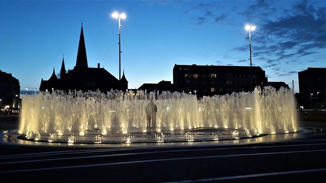 Aarhus city centre. We’re looking at a fountain embedded directly in the ground in the harbour promenade. Thin two-metre jets of water shoot up at random intervals in big spirals. As I took this photo, all the jets are in full swish. The sun has just set, the sky is a mid-Summer light night-blue. The skyline is the cathedral spire and turrets and spires seen in silhouette. The ground at our feet is black in darkness. By contrast, lights in the fountain make the jets dazzle and sparkle with a creamy white colour. There is space between the jet spirals. Adults and children love playing in them, day and night. Inside the spirals of fountains at this moment, the silhouette of a couple standing romantically close together.