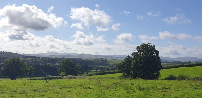 A view across fields and woods to 2 distant peaks of the western edge of the Bannau Brycheniog national Park. The sky us blue with white clouds