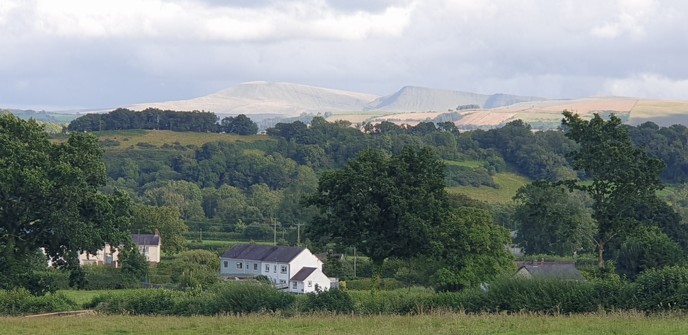 On the foreground some fields then 2 houses and another hidden behind trees. Then there are a lot of trees and some fields and beyond that 3 peaks of the Bannau Caerfyrddin bathed in sunshine with a cloudy grey sky above