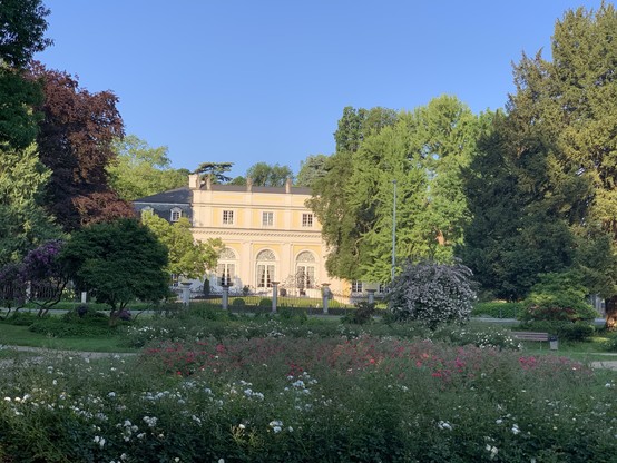 Elegant mansion surrounded by lush green trees and blooming gardens under a clear blue sky.