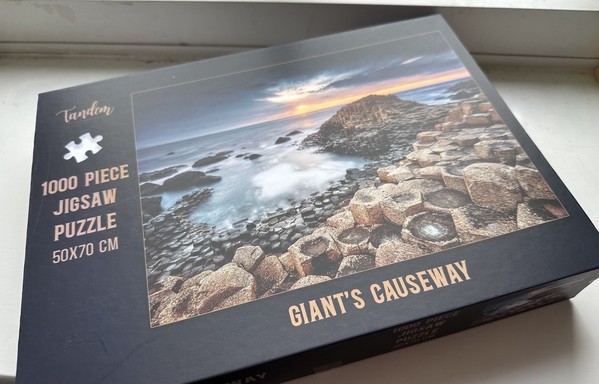 Jigsaw box with a gorgeous sunset photo of the Giant’s Causeway on the front. In the foreground are some of the hexagonal basalt pillars making up the causeway, snaking towards the mound at the end, which is largely surrounded by the foaming tide. The sun is setting into a calm flat sea at the horizon and, above, the light has caught the underside of the clouds which are otherwise in shadow