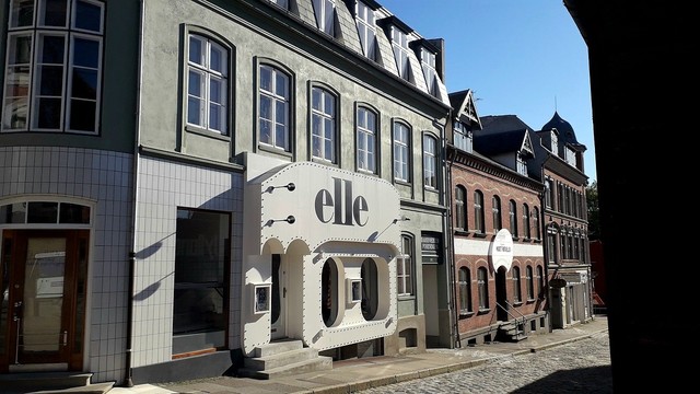 Looking along a street in the Yikes My Childhood Is In A Museum section of Den Gamle By, the Old Town open-air museum in Aarhus. The 1970s section. One shop stands out. It's the original facade of a women's fashion boutique. The shop front is shaped like a gigantic white cassette tape with the windows like the holes where you'd stick a pen in to try and wind loose tape back on. The shop name Elle is on the 'tape' in giant black letters. It's a funky, clever design that catches the eye, even if you aren't old enough to recognise the shape.
