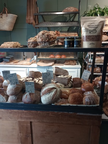 The interior of a small bakery with a glass and wood show cabinet laden with various loaves and buns and cakes. There's a packet of granola on teh top, and in the background, trays of freshly baked buns are visible. 