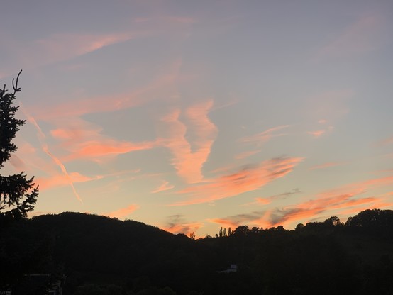 Sunset with pink and orange clouds over a dark landscape with trees and hills.