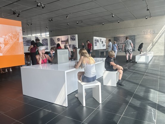 Our children at the Topograpie des Terrors exhibition in Berlin. Large hall, display posters in background, white table and chairs with boy and girl sitting on them. 