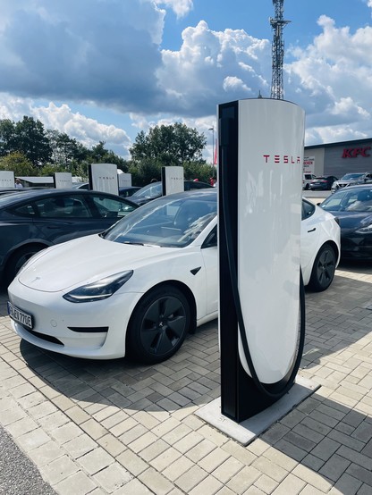 A white Tesla car parked at a Tesla Supercharger station in white and black with several other cars and a KFC building in the background under a cloudy sky.