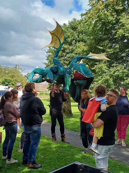 Puppet dragon with three puppeteers on the village green. Small crowd assemble. Big wings!