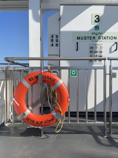 A muster station on a white ship with signage and a bright orange life ring labeled “MS NORDERAUF