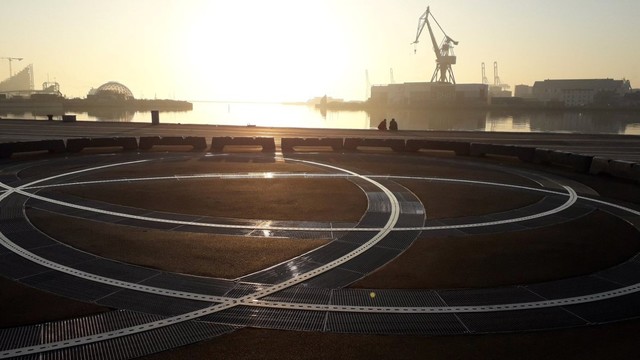 Aarhus harbour, a misty dawn a few years ago. The concrete promenade stretches out for several hundred metres at our feet. Embedded in it, spirals of metal that are the source of jets of water when a fountain is activated here. They are gleaming silver in the dawn light. Ahead, the sea and pier and sky look yellow-white in washed-out backlight. We can see the geodome and harbour cranes as if through a mist. By the water, the silhouettes of two people sitting close together, legs dangling over the parapet edge. The scene feels to me like a sepia photo. From the past. It looks quiet. It looks as if the two people are also not talking, just enjoying a quiet dawn together.  