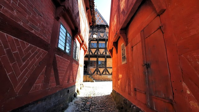 A narrow lane between two medieval houses in Den Gamle By Open-Air museum, Aarhus, looking ahead to a third historic house on the street. The wood and brickwork on the houses is red and orange, giving them warmth and depth. They are criss-crossed with timbered planks. They have multi-paned windows. The street is cobblestoned. A lovely old lamp with a triangular top juts out high up on one of them, hanging on a curly ornate bracket. There is nothing to the scene. Objectively, it is dull. I just feel it also has a sense of quiet and peace about it. As sense of taking a moment away from the world, here in this old lane where people walked back in the 15th and 16th centuries 