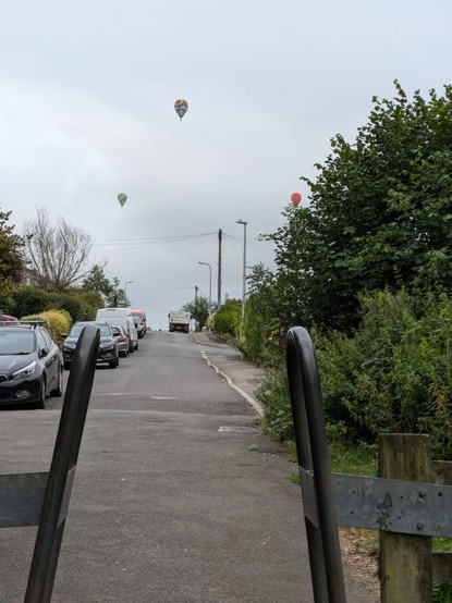 Three hot air balloons above a quiet suburban street leading away from the end of a bike path.