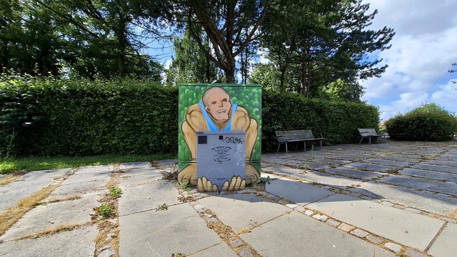 A long wall, benches, trees, wide pavement by an Aarhus cemetery. In front of us: a big utility box with a small utility box at its base. The big box has been painted with an image of a stereotypical strongman, a white man in a blue vest in front of green leaves, in a squat, his muscular arms and big feet showing on either side of the little box. The little box is not painted, it’s just the original grey metal, except that the fingers of the strongman that have been painted on the bottom, as if grabbing it from behind and underneath. It makes it look as if he is straining to lift the little box. On the little box, someone has graffitied the words: ‘No human being is useless or unproductive. There are only people whose contributions under capitalism are not valued.’ Someone else (I assume) has painted a stick-figure of a white chicken with a confused expression beside this. This juxtaposition left me with an expression like a confused chicken too. Now follows same context description for all photos in this thread, if you want to skip. A council utility box for electrical and technical stuff, about half a metre by half a metre in size, normally grey metal box, typically dirty and covered in graffiti and stickers. They have been transformed all across the city in recent weeks, painted on all four sides and on top with images that reflect something in the immediate neighbourhood. Artist signature: Johan Wiklund.