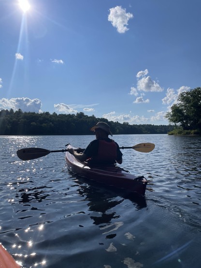 A person kayaking on a calm lake under a clear blue sky with a few clouds. The sun is bright, creating reflections on the water. Trees line the far shore.