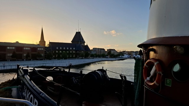 Aarhus harbour. Taken just after sunset, after 10pm, on the summer solstice, so the sky remains light and blue. In the midsummer night light, the sea is silver-blue and the promenade gleams white. Right and along the bottom of the image: the prow and wheelhouse of the dark blue old trawler Jakob, a red-and-white lifebelt and green ropes hanging on its red-and-white funnel. Framed above the boat, the city skyline with turrets and the cathedral spire visible, not even silhouettes because of the white-nights light. A glow of fiery orange is seen behind the spire, the remains of the day, the remains of the sun. 
