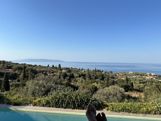 View over olive groves and scattered buildings to the Ionian Sea and the island of Zakynthos on the right, a pair of crossed feet in front of a swimming pool in the foreground 